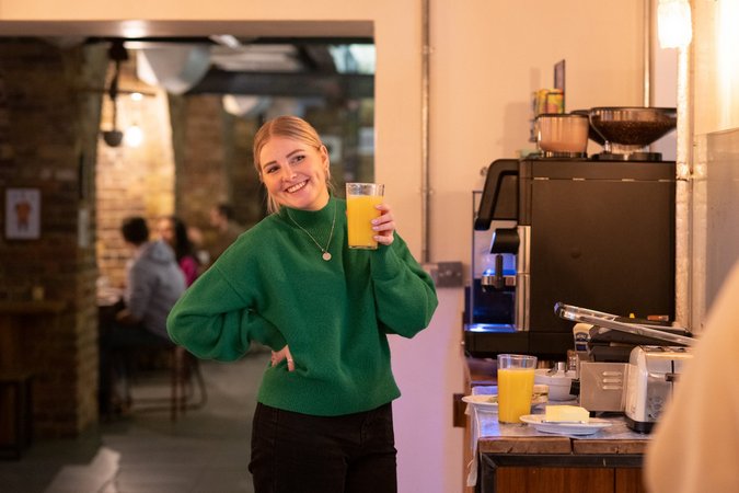 A Girl raises a glass of fresh orange juice with a bright smile, symbolizing the vibrant and refreshing start to the day offered at the hostel.